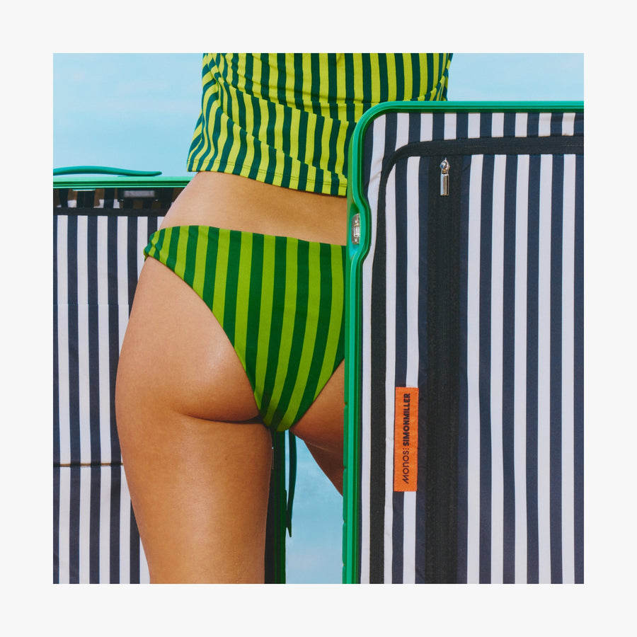 Coastal Blue | Close-up of a woman in a green striped swimsuit standing beside an open Jungle Green Hybrid suitcase, revealing its interior Poplin Stripe Black interior