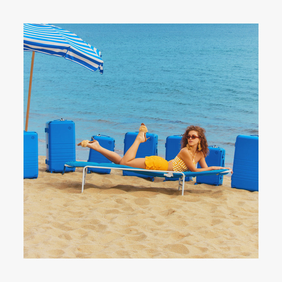 Jungle Green | Woman lounging on a beach chair under a striped umbrella, with Coastal Blue Hybrid suitcases arranged behind her on the sand by the ocean
