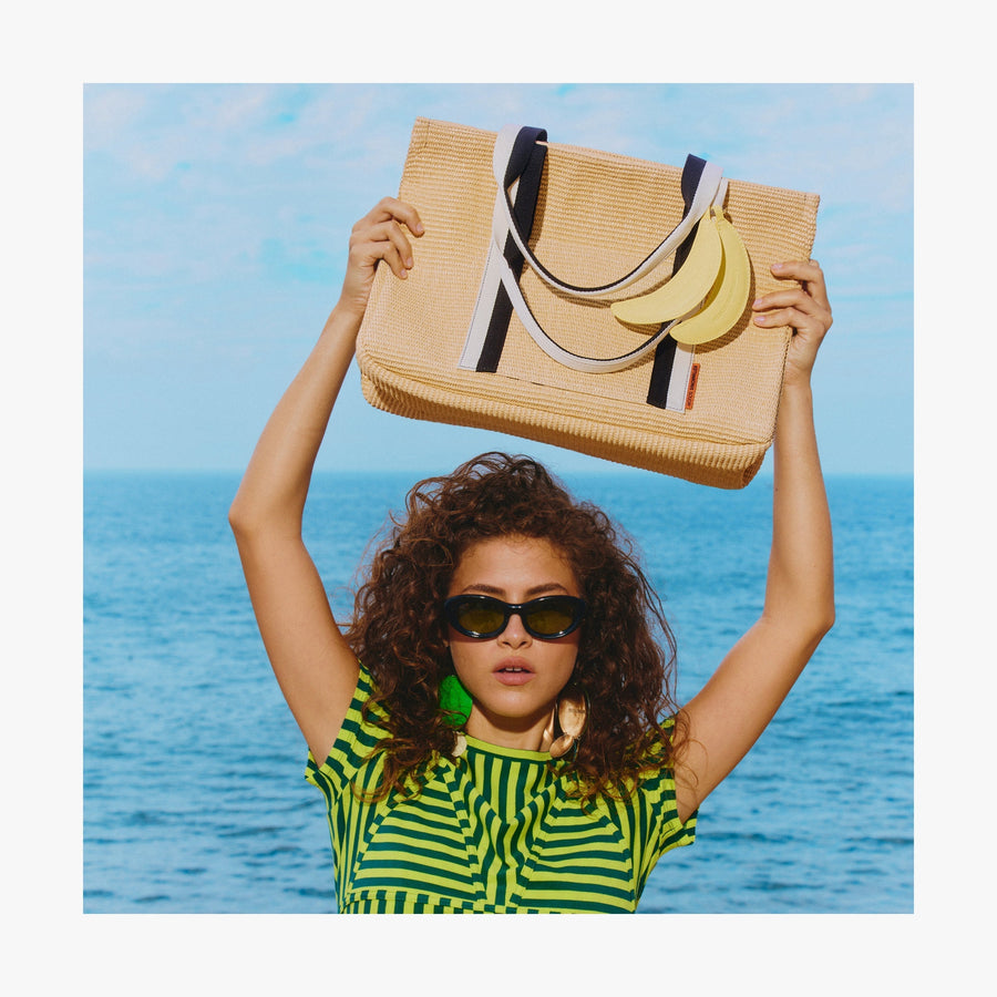 Natural | Woman holding a  Canopy Tote with the Banana Charm above her head while standing by the ocean