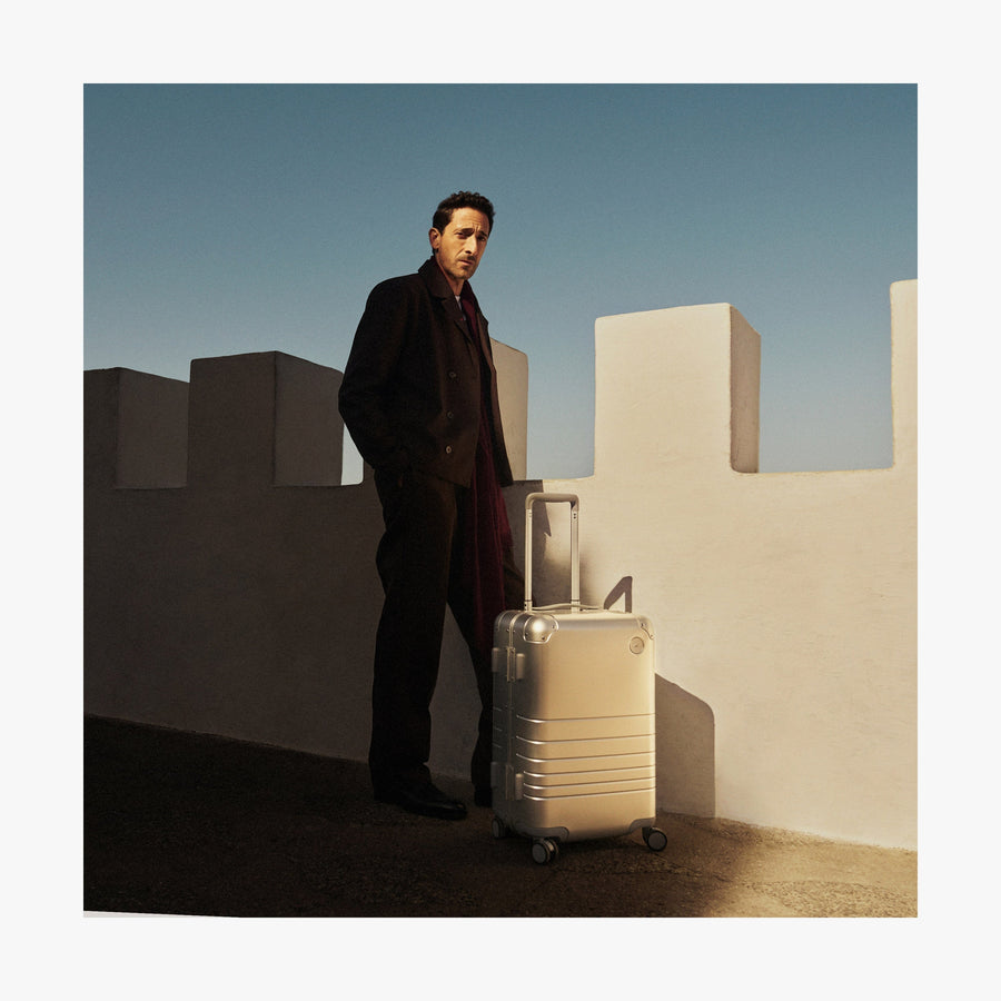 Caviar Black | Adrien Brody wearing a dark coat and red scarf stands beside a white fortress-like wall, with a Monos aluminum carry-on plus in aspen silver in the sunlight. The deep blue sky contrasts with the shadows.