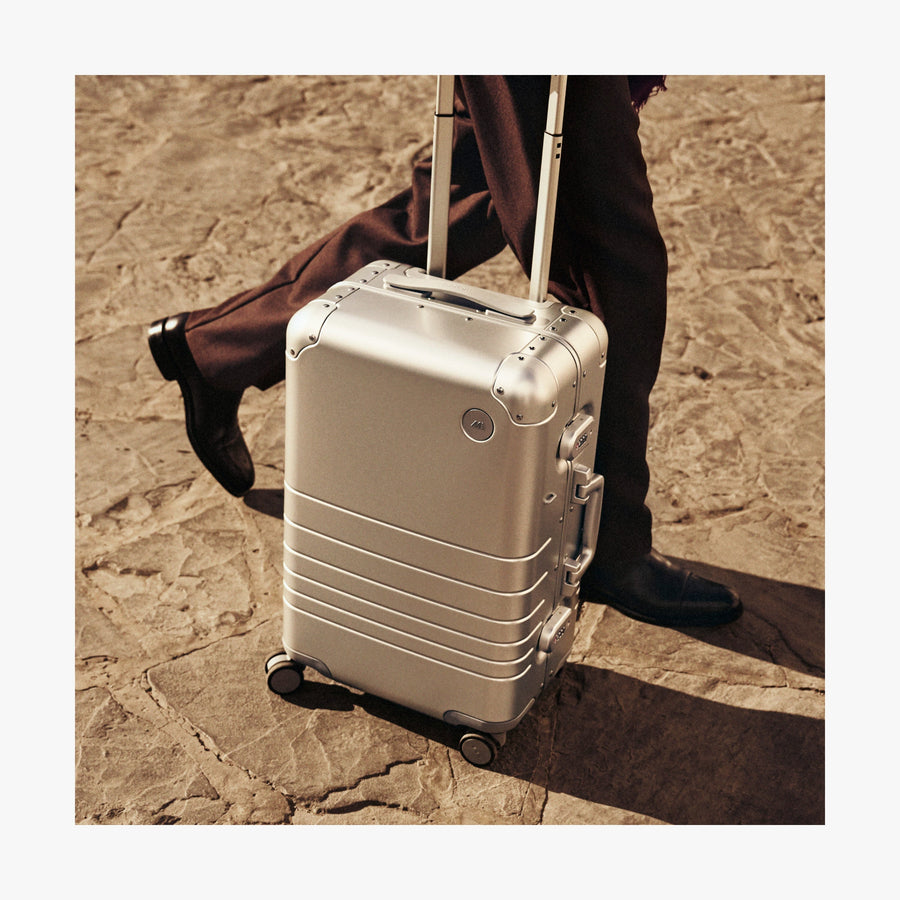 Caviar Black | A close-up of a Monos aluminum carry-on plus suitcase in aspen silver. A person wearing dark brown trousers and polished black shoes walks alongside it on a textured stone pavement, with warm sunlight casting soft shadows.