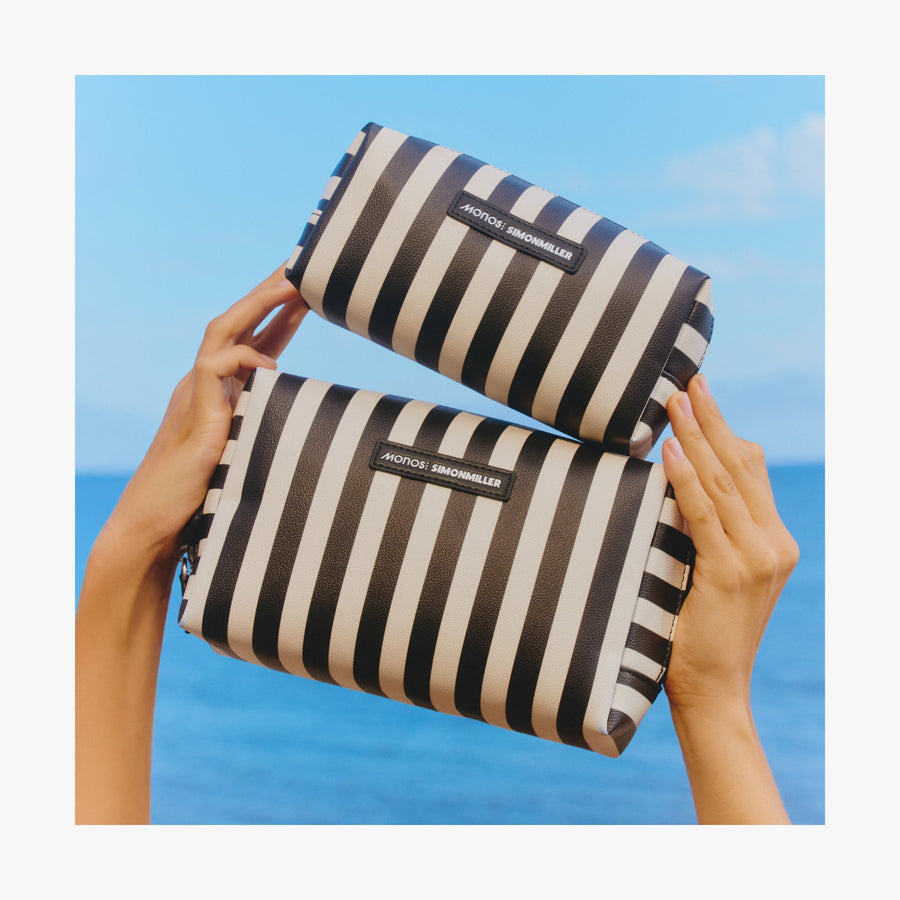 Small / Poplin Stripe Black | Two hands holding up the two sizes od Metro Toiletry Cases in Poplin Stripe Black against a bright ocean background
