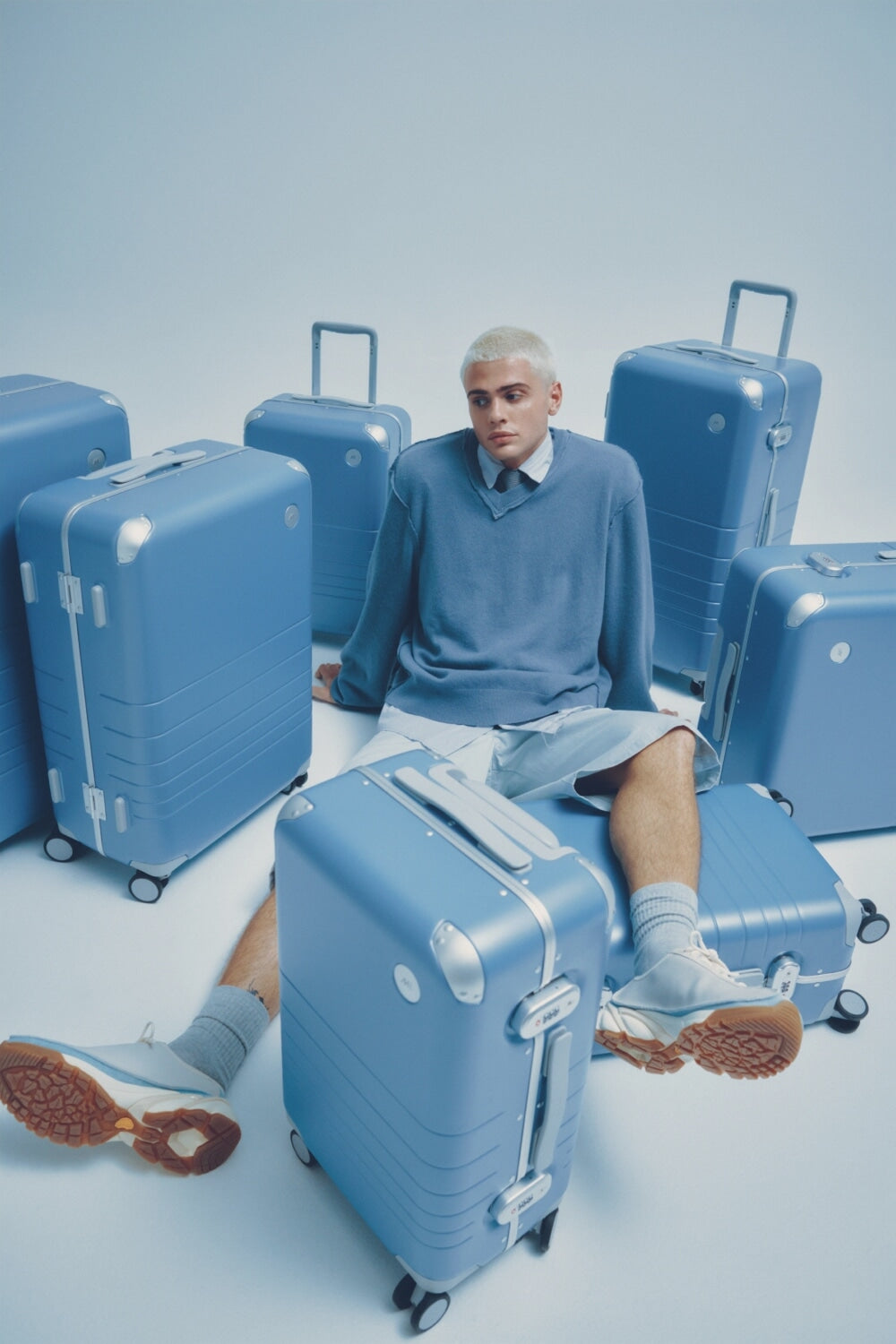 A model in a blue sweater sits among various sizes of Hybrid luggage in Elemental Blue on a light blue background.