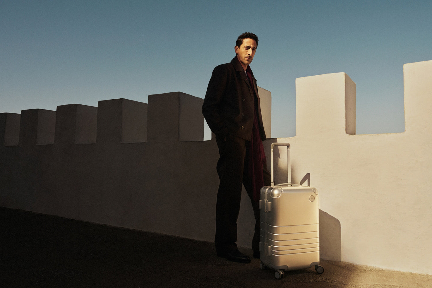 Adrien Brody wearing a dark coat and red scarf stands beside a white fortress-like wall, with a Monos aluminum carry-on plus in aspen silver in the sunlight. The deep blue sky contrasts with the shadows.