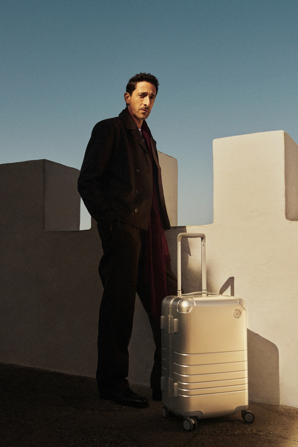 Adrien Brody wearing a dark coat and red scarf stands beside a white fortress-like wall, with a Monos aluminum carry-on plus in aspen silver in the sunlight. The deep blue sky contrasts with the shadows.