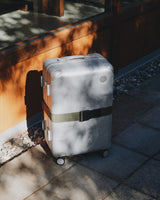 Hybrid Carry-On in Silver with Luggage Strap in Olive Green under soft sunlight.