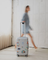 The Monos Hybrid Carry-On in silver, decorated with Destination Stickers, rolls across a sunlit floor beside a blurred figure in blue.