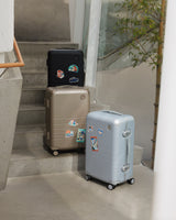 Three Monos Hybrid Carry-On suitcases in Obsidian, Champagne, and Silver stand on concrete steps, each personalized with Destination Stickers.