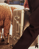 A close-up of the man’s hand gripping the handle of a Monos aluminum suitcase in aspen silver, walking past a woolly sheep in a rustic outdoor setting.