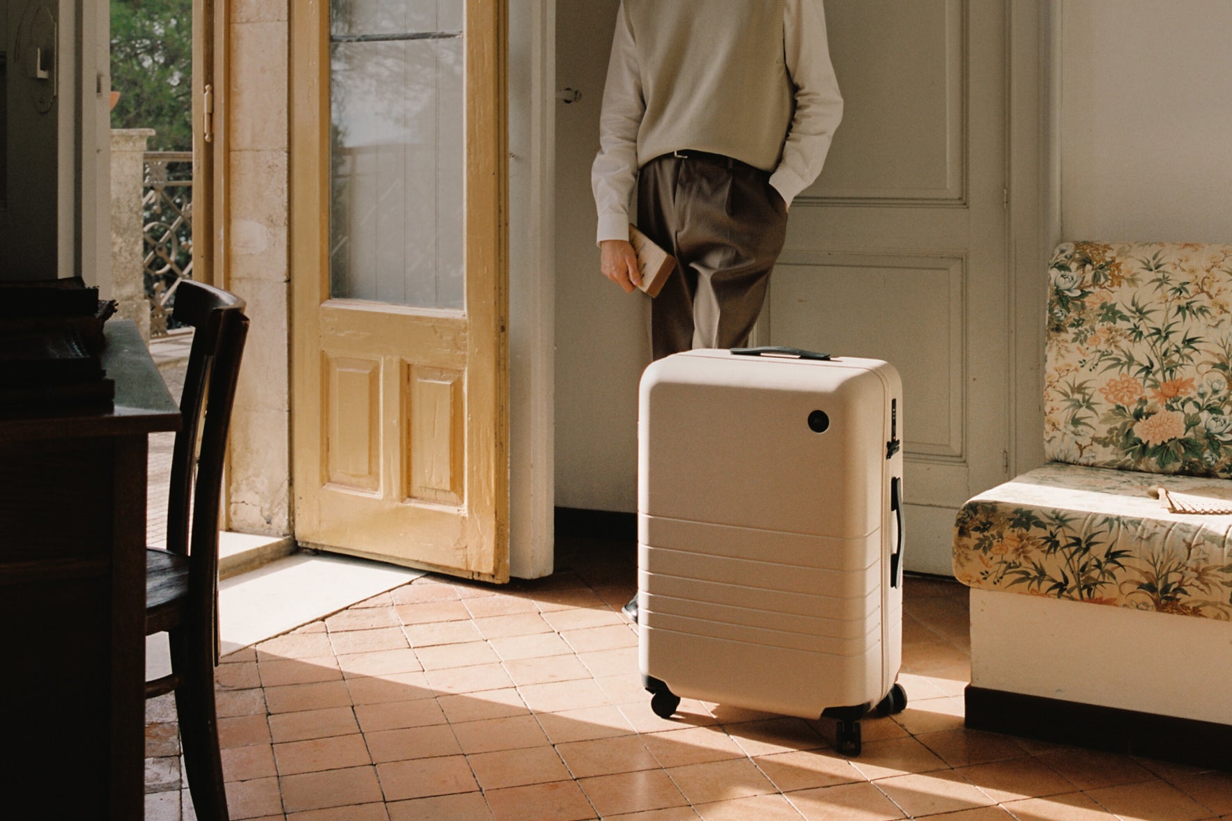 Person holding a white suitcase in a room with a wooden door and floral-patterned chair.